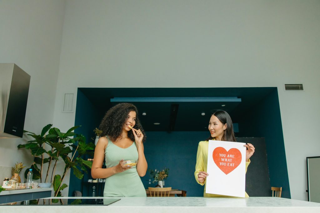 Client enjoys a healthy snack while a personal nutritionist holds a "You Are What You Eat" sign in a modern kitchen.